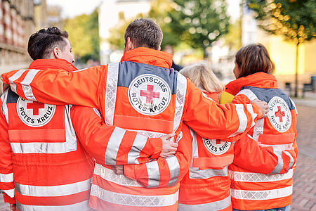 Unser Symbolfoto zeigt zwei Rotkreuzler und zwei Rotkreuzlerinnen, die mit dem Rücken nebeneinander zur Kamera stehen und eine Reihe bilden. 